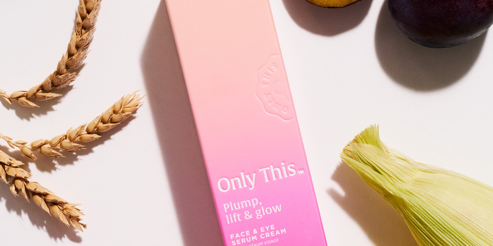 Pink skincare product box labeled 'Only This Plump, Lift & Glow' on a white surface with corn, plums, and flowers.